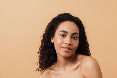 Shirtless black woman with wavy hair posing and looking at camera isolated over beige background