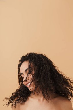Shirtless black woman laughing while making fun with her hair isolated over beige background