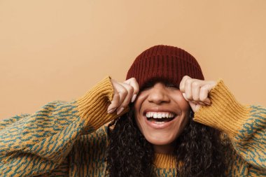 Young black woman laughing while making fun with hat isolated over beige background