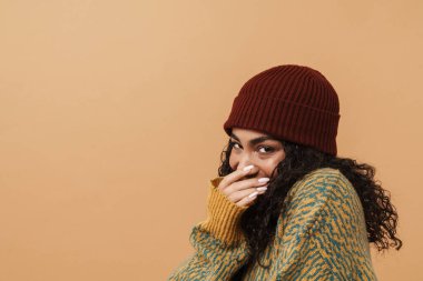 Young black woman wearing hat laughing and covering her mouth isolated over beige background