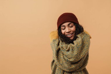 Young black woman wearing hat laughing and hugging herself isolated over beige background