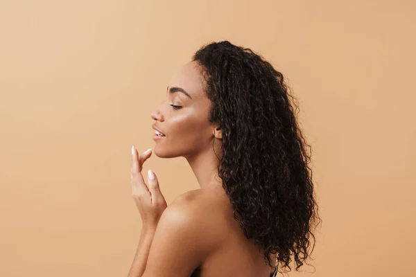 Shirtless black woman with wavy hair touching her facial skin isolated over beige background