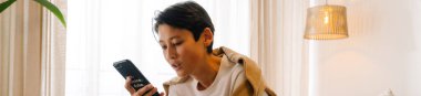 Asian boy using mobile phone while sitting on bed at home
