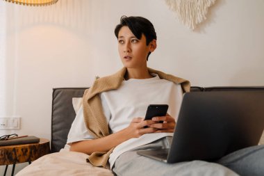 Asian boy using cellphone and working with laptop while resting on bed at home