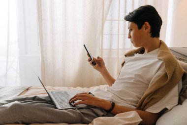 Asian boy using cellphone and working with laptop while resting on bed at home