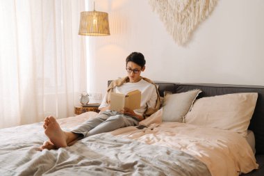 Asian boy in eyeglasses reading book while resting on bed at home