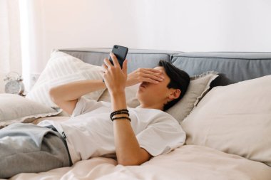Asian tired boy using mobile phone while resting on bed at home