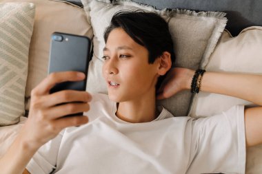 Asian boy using mobile phone while resting on bed at home