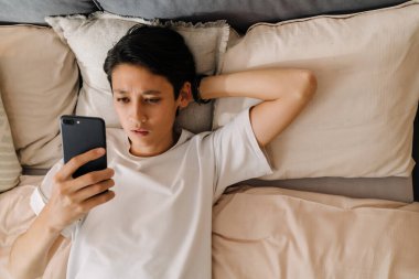 Asian boy using mobile phone while resting on bed at home