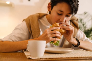 Asian brunette boy eating sandwich while sitting at desk in kitchen