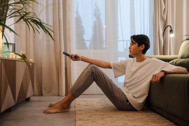 Asian boy using remote control and watching tv while sitting on floor at home