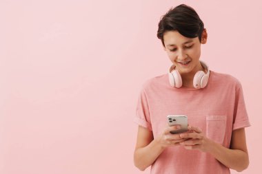 Asian boy wearing t-shirt smiling while using mobile phone isolated over pink background