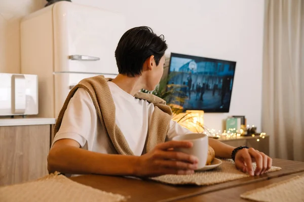 Asian brunette boy watching tv while having breakfast in kitchen