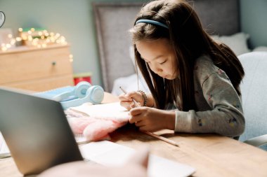 Asyalı kız evde otururken dizüstü bilgisayarla ödev yapıyor.