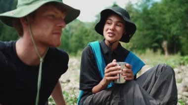 Yakışıklı, çok uluslu bir turist çifti dağlarda nehir kenarında çay içerek konuşuyor.