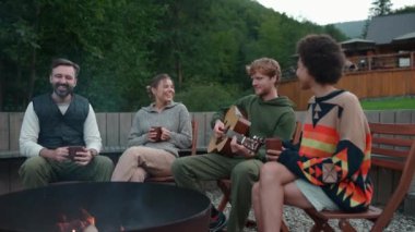 Dağlarda ateşin etrafında oturmuş gitar çalan gülen çok uluslu arkadaşlar.