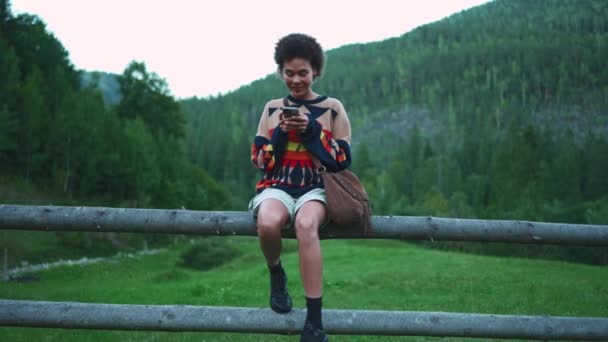 Dağlardaki Ahşap Bir Çitte Otururken Telefonda Yazan Gülümseyen Afrikalı Kadın — Stok video