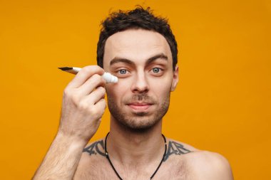 Orta yaşlı beyaz bir adam turuncu arka planda yüzüne nemlendirici krem sürüyor.