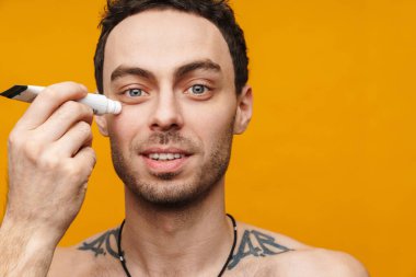 Orta yaşlı beyaz bir adam turuncu arka planda yüzüne nemlendirici krem sürüyor.