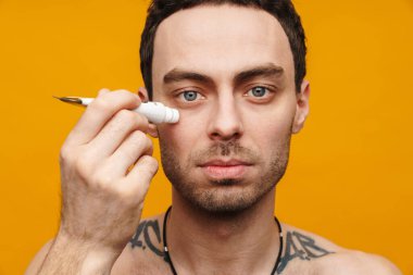 Orta yaşlı beyaz bir adam turuncu arka planda yüzüne nemlendirici krem sürüyor.