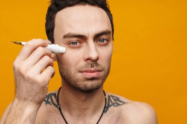 Orta yaşlı beyaz bir adam turuncu arka planda yüzüne nemlendirici krem sürüyor.