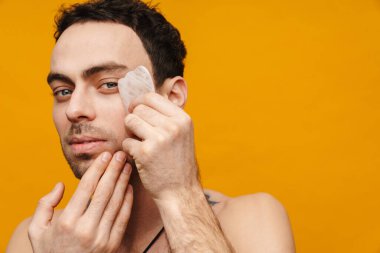 Esmer, orta yaşlı bir adam yüzüne sarı arka planda kozmetik bir silindirle masaj yapıyor.