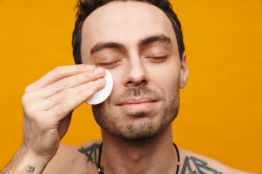 Üstsüz gülümseyen orta yaşlı esmer bir adamın yüzünü sarı arka planda pamukla temizlerken.