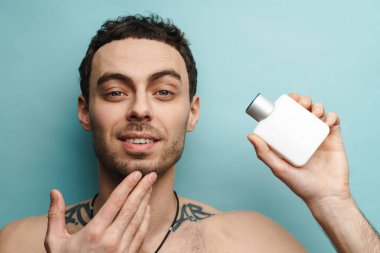 Mavi duvarda izole edilmiş parfüm şişesini gösteren gömleksiz beyaz adam kameraya bakıyor.