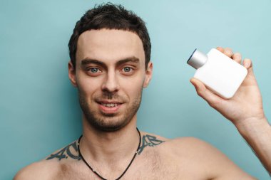 Mavi duvarın üzerinde parfüm tutarken gülümseyen gömleksiz beyaz adam.