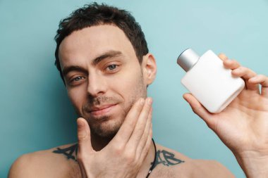 Mavi duvarın üzerinde parfüm tutarken gülümseyen gömleksiz beyaz adam.