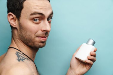 Mavi duvarın üzerinde parfüm tutarken gülümseyen gömleksiz beyaz adam.