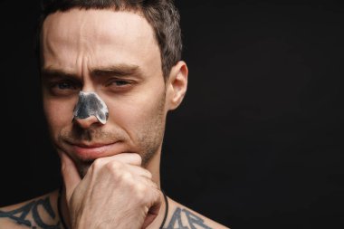 Orta yaşlı, beyaz gömleksiz bir adam siyah duvar arka planında burun temizleme bandı kullanıyor.