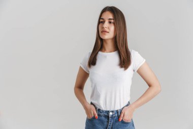 Tişörtlü, uzun esmer saçlı, gri duvarın üzerinde duran, cepleri boş genç beyaz bir kadının portresi.