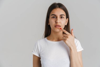 Kafası karışmış, beyaz tişörtlü, uzun esmer bir kadının portresi. Gri duvarın arkasında durmuş dudaklarını işaret ediyor.