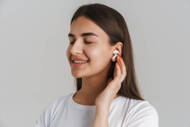 Uzun esmer saçlı, gri duvar arkasında dikilmiş, kablosuz kulaklıkla müzik dinleyen genç, mutlu beyaz bir kadının portresi.