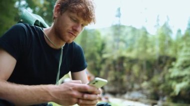 Güzel turist kızıl saçlı adam dağlarda çadırın yanında otururken cep telefonuyla mesajlaşıyor.
