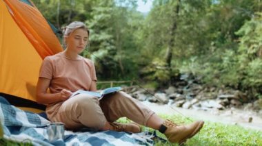 Dağlarda çadırın yanında oturmuş kitap okuyan mutlu turist kadın.