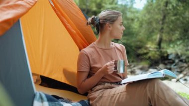 Kitap okuyan ve dağlarda çadır başında çay içen pozitif turist kadın.