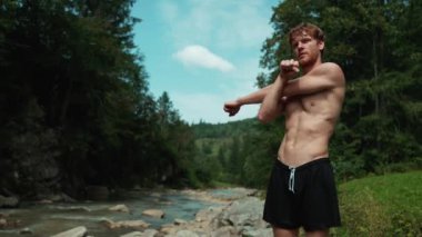 Dağlarda nehir kenarında esneme egzersizi yapan konsantre kızıl saçlı adam.