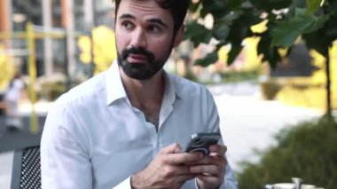 Beyaz tişörtlü gülümseyen esmer adam dışarıda bankta cep telefonuna yazı yazıyor.