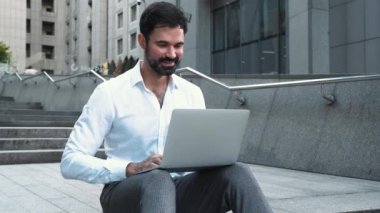 Beyaz tişörtlü gülümseyen esmer adam dışarıda dizüstü bilgisayarla çalışıyor.