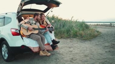 Mutlu sarışın çift gitar çalıyor ve arabanın bagajında şarkı söylüyor.