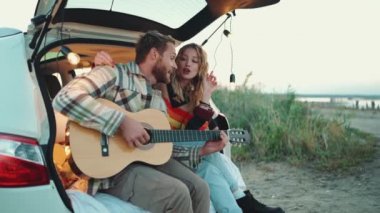 Güzel sarışın genç bir çift arabanın bagajında gitar çalıp şarkı söylüyor.