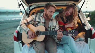 Arabanın bagajında gitar çalıp dans eden genç, pozitif sarışın bir çift.