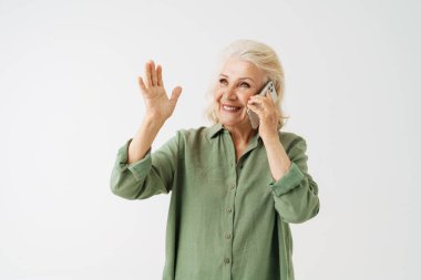 Beyaz arka plan üzerinden izole edilmiş cep telefonuyla konuşurken tişörtlü yaşlı bir kadın el kol hareketi yapıyor.