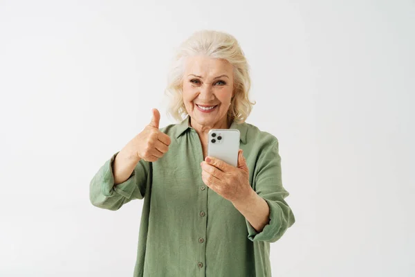 在使用白色背景隔离的手机时 灰色的老年妇女露出大拇指 — 图库照片