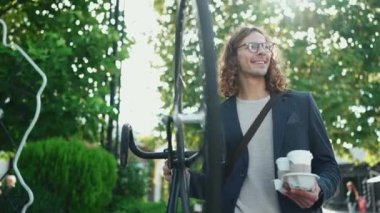 Ceket ve gözlük takan, gülümseyen kıvırcık saçlı adam parkta bisiklet ve bir fincan kahveyle ayakta duruyor.