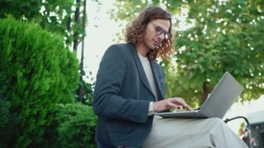 Kendine güvenen kıvırcık saçlı, ceketli adam dışarıda dizüstü bilgisayarla çalışıyor.