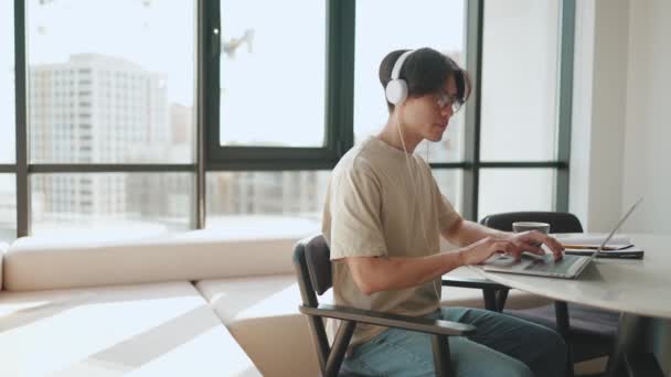 自信を持ってアジアのブルネットの男は自宅のキッチンでヘッドフォンでラップトップで作業 — ストック動画