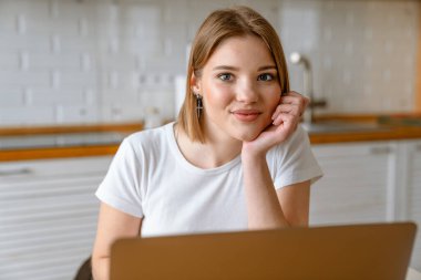 Gülümseyen sarışın beyaz kadın mutfak masasında dizüstü bilgisayarla oturuyor.